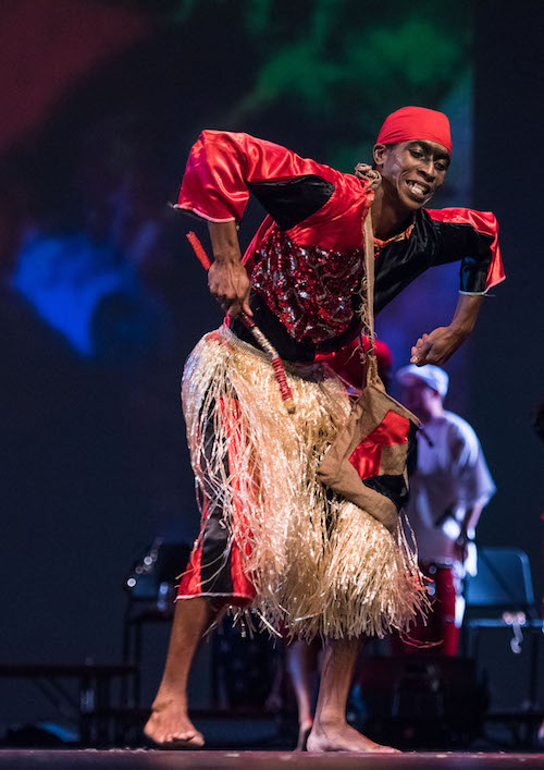 A man in a red and black costume and grass skirt. He hunches his over while his arms are bent the elbows with his hands facing down
